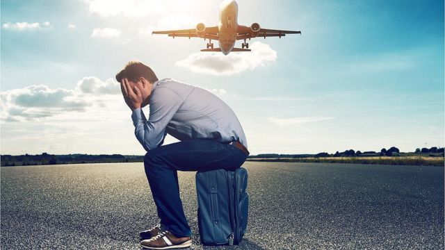 Hombre infeliz sentado sobre su maleta en la pista mientras avión vuela.