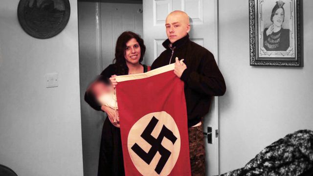 Claudia Patatas com o filho no colo ao lado de Adam Thomas, que segura uma bandeira nazista