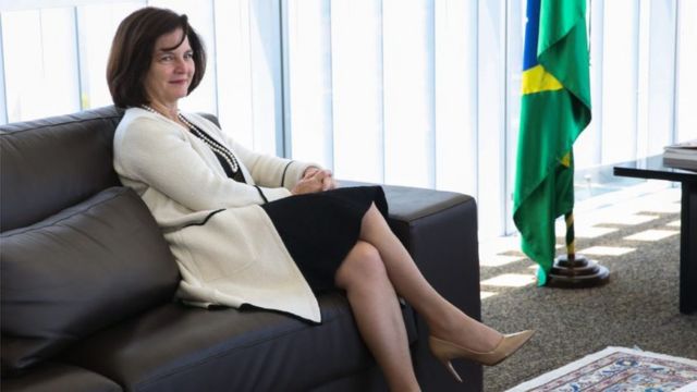 A procuradora Raquel Dodge sentada em um sofá escuro ao lado de uma bandeira do Brasil