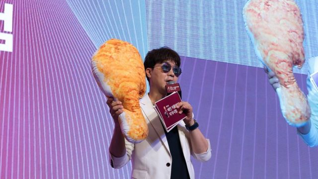 Famous Korean TV presenter speaks into a microphone on stage as he holds a giant cushion shaped like a fried chicken drumstick