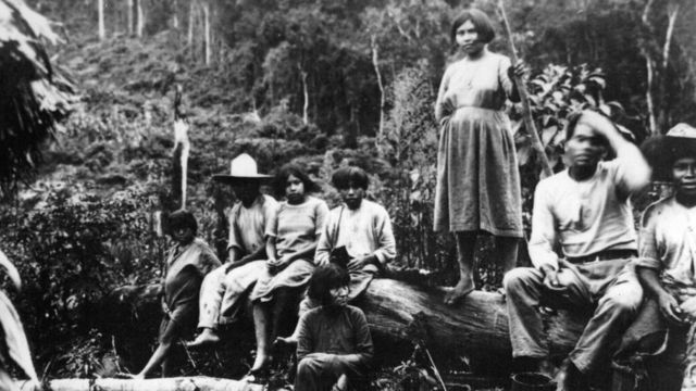 Indígenas sentados em pedaço de madeira