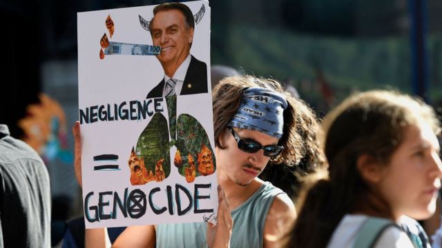Ativista segura cartaz com uma foto do presidente Jair Bolsonaro e de florestas em chamas, com as palavras 'negligência' e 'genocídio' em inglês. Londres, agosto de 2019