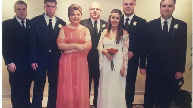 Luiz (no centro) ao lado da esposa, de filhos e de nora posam para foto em casamento. Homens de terno, esposa com vestido rosa e nora está de noiva.