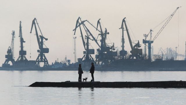 Fotografia mostra silhueta de casal com cachorro em frente aos guindastes do porto de Mariupol
