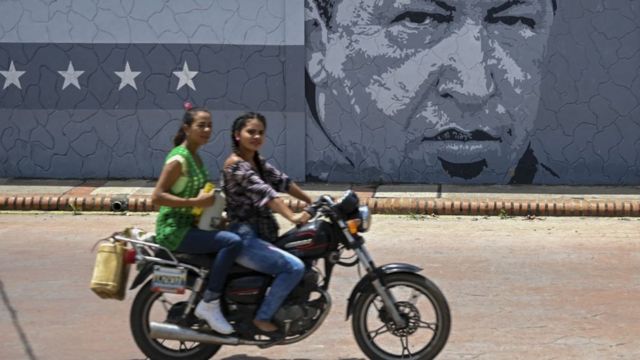 Two women ride a motorcycle past a mural with the face of Hugo Chávez in Barinas.