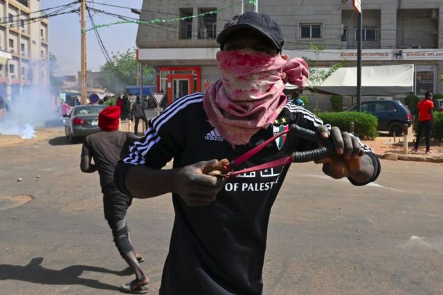 A masked man readies a slingshot on a street with protesters and tear gas behind him.