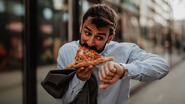 Un hombre comiendo un trozo de pizza toda prisa porque tiene que volver al trabajo.