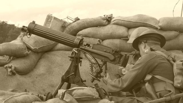 Fotografia preto e branco de dois soldados manipulando armamento pesado atrás de uma trincheira