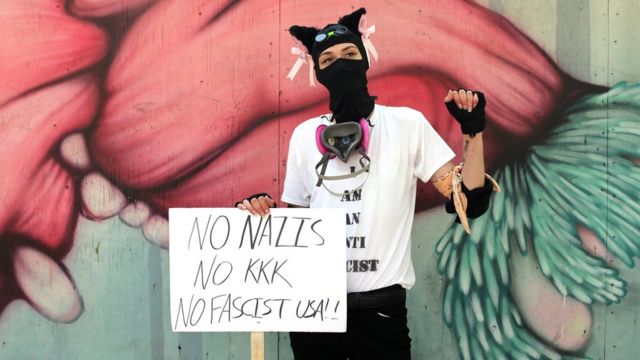 Mulher aparece em frente a muro grafitado usando uma máscara, com um braço levantado e um cartaz dizendo: "Nazistas não"