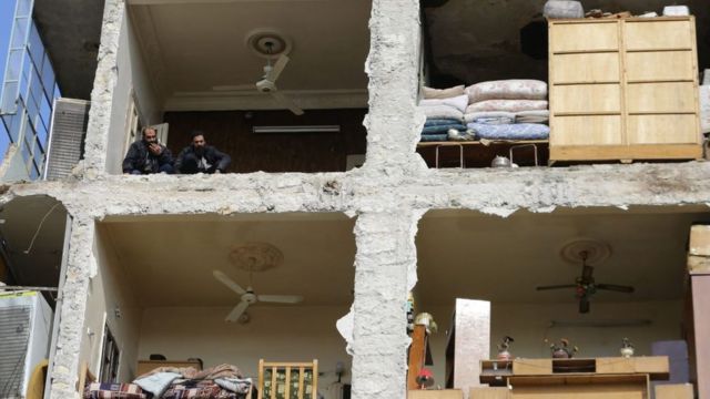 Dos hombres observan desde el último piso de un edificio dañado mientras los equipos de rescate buscan víctimas y sobrevivientes atrapados bajo los escombros en la ciudad de Alepo, en el norte de Siria.