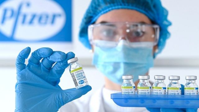 Photo of a healthcare professional wearing a cap, gloves, mask, glasses and apron holding a tray with vaccine vials