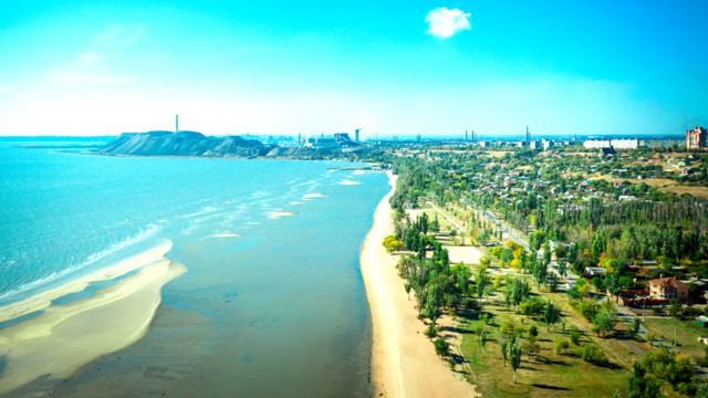 Fotografia mostra Paisagem da costa de Mariupol antes da guerra, praia, mar azul e muito verde entre os prédios da cidade