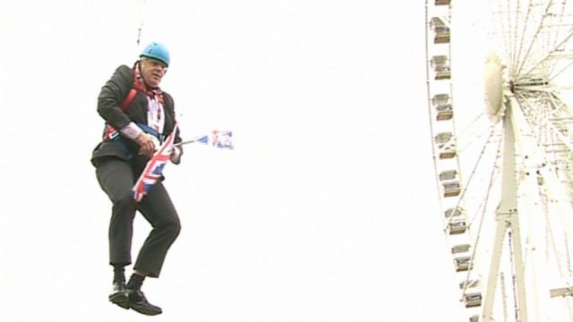 بوريس جونسون متدليا من سلك مضغوط بينما يحمل أعلام المملكة المتحدة ويرتدي بدلة رسمية