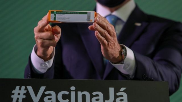 De máscara, João Doria mostra embalagem da Coronavac, com palanque em baixo exibindo a palavra #VacinaJá