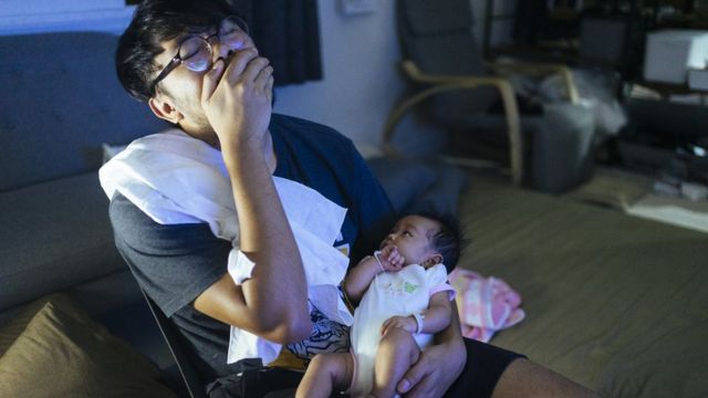 Padre bostezando con su bebé despierto en brazos durante la noche.
