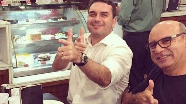 Sentados, Flávio Bolsonaro e Queiroz fazem sinal de arma para foto de dentro de restaurante