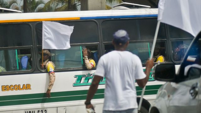 Combatientes de las FARC saludan desde un autobús a un hombre vestido de civil que sostiene una bandera blanca.
