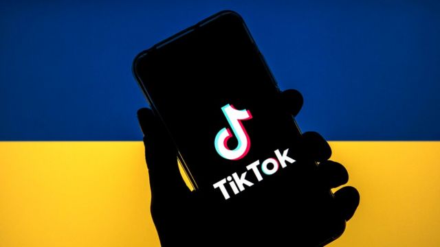 A TikTok logo displayed on a smartphone with a flag of Ukraine in the background.