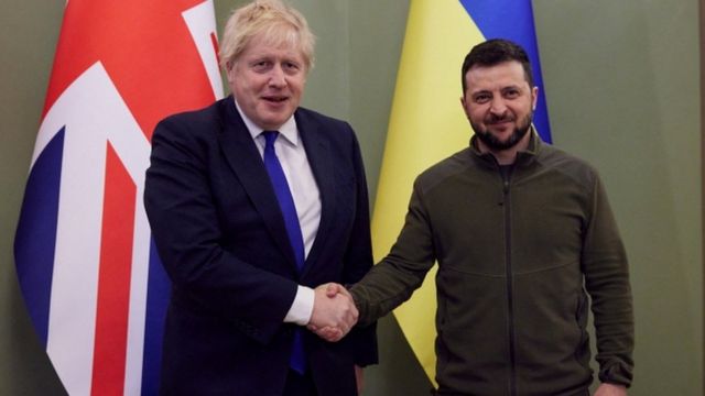 Boris Johnson e Volodymyr Zelensky apertão a mão um do outro em frente às bandeiras de seus respectivos países