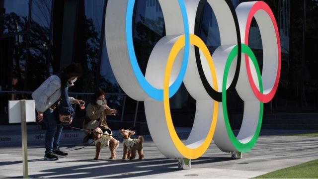 Pessoas interagem em um banco público atrás do símbolo da Olimpíada