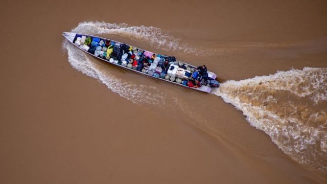 Imagem aérea mostra barco carregado de barris e pessoas em água marrom do rio