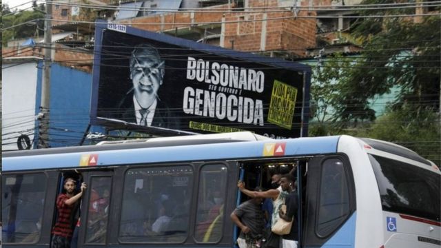Os passageiros viajam em um ônibus público lotado, passando por uma painel contra o presidente Jair Bolsonaro