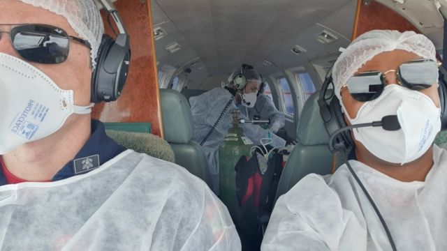 Pilotos aparecem à frente com paciente com oxigênio medicinal ao fundo dentro de aeronave