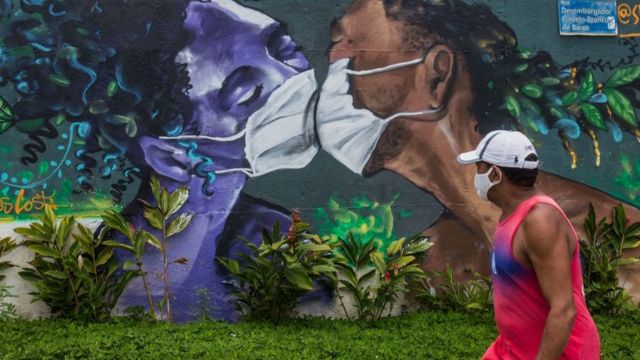 Un hombre con mascarilla camina frente a un mural en el que dos personas pintadas con mascarillas se besan.