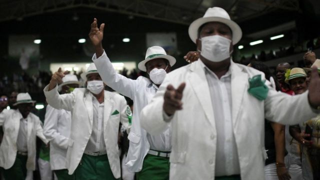 Homens com trajes típicos de sambistas aparecem gesticulando enquanto cantam com máscara