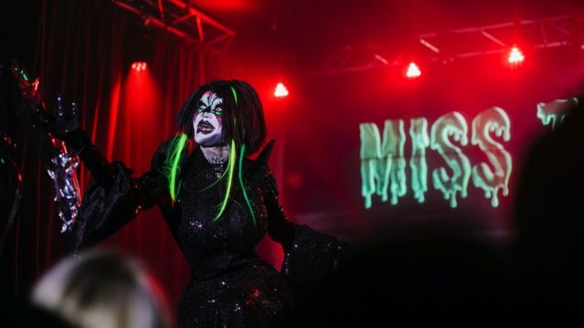 Danya en el escenario con un vestido ajustado negro con maquillaje blanco y extensiones de cabello de neón.