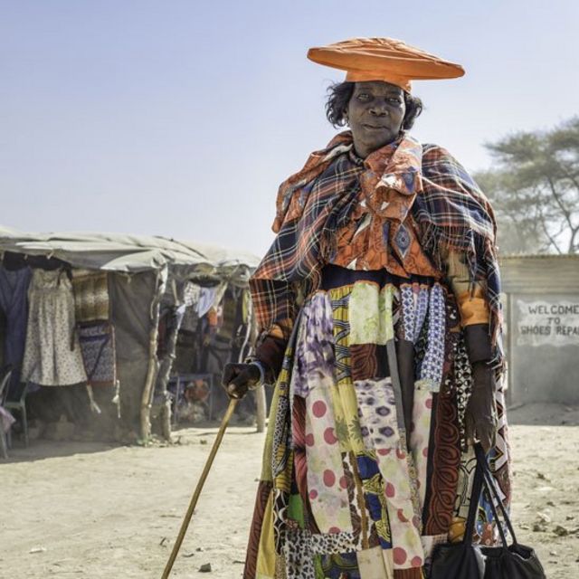Mujer Herero con un bastón en una mano y su cartera en otra, frente a una casa precaria hecha con palos y telas