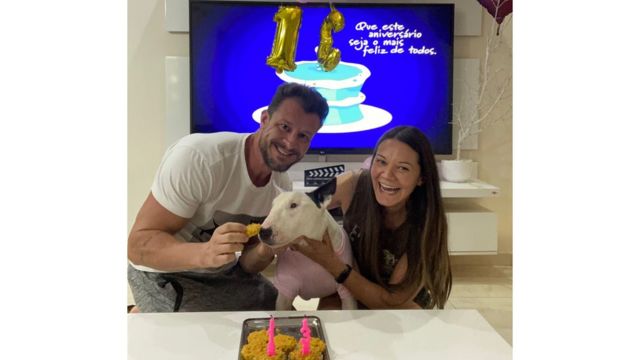 Casal sorrindo com cadela no meio, em frente a bolo de aniversário
