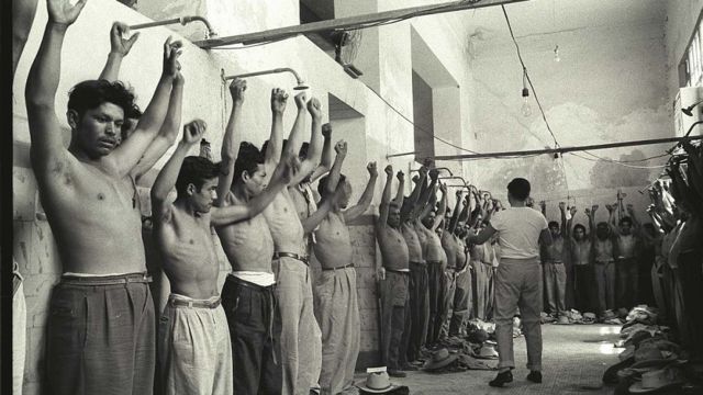 Un grupo de trabajadores del programa Bracero alzan los brazos y están alineados contra la pared mientras son inspeccionados en una habitación del Centro de Procesamiento en Monterrey, México.