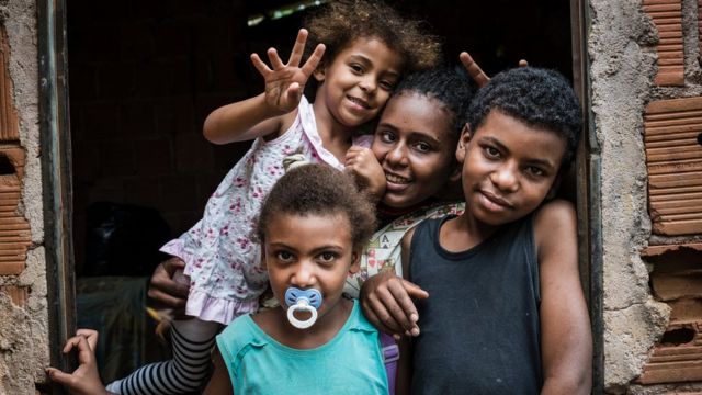 Um menino e três meninas negras no batente de uma porta em casa de tijolos sem acabamento