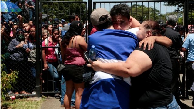 Una mujer se abraza con dos hombres frente a una multitud detras de las rejas en Managua.