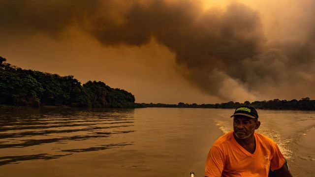 Pescador anda em barco com grande nuvem de fumaça ao fundo