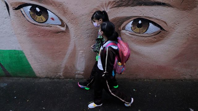 Niñas yendo al colegio pasando frente a un mural que muestra los ojos de un niño