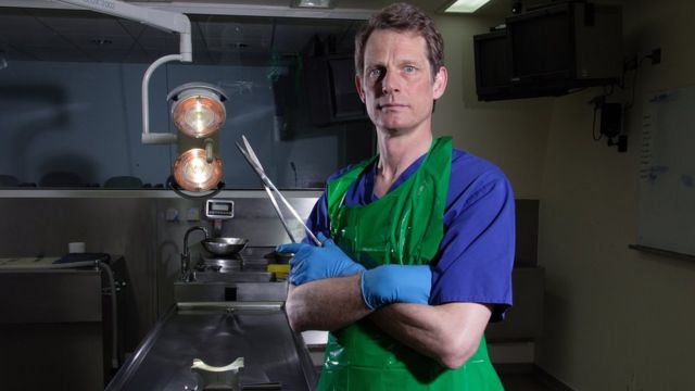 Dr Richard Shepherd dentro de laboratório, com ferramenta em forma de tesoura nas mãos e jaleco