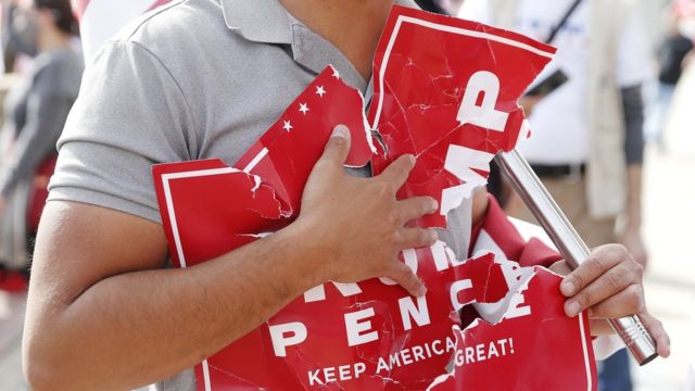 Un hombre sostiene un letrero roto que dice "Trump y Pence".