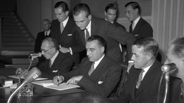 Foto em preto e branco de um grupo de homens reunidos em torno de uma mesa assinando um documento