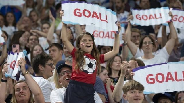 Niñas y mujeres que son hinchas de fútbol sostienen carteles que dicen "¡Gol!"durante un partido de Inglaterra en la Eurocopa Femenina 2022