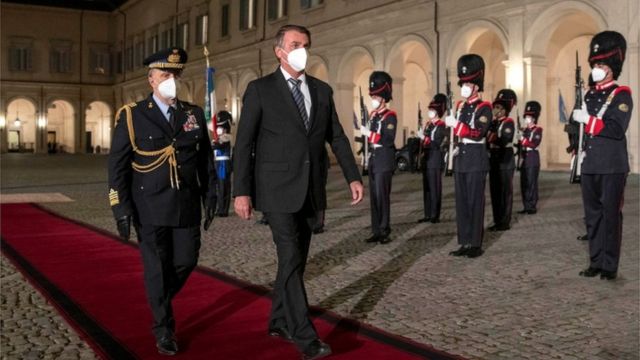 Bolsonaro caminha de máscara em tapete vermelho, rodeado por militares