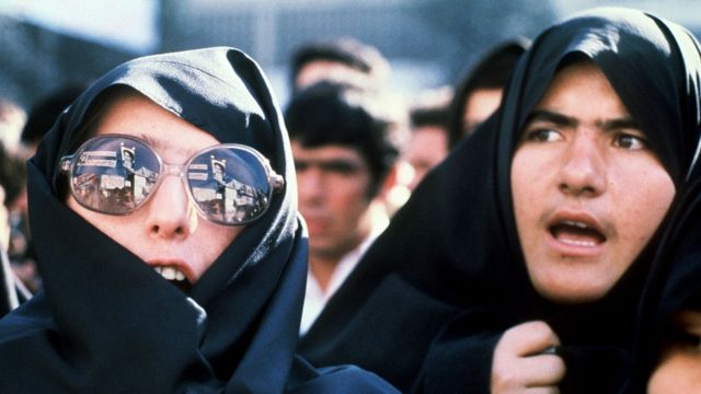 Iranke pokrivenih glava - jedna od njih nosi naočare za sunce na kojima se reflektuju slogani i portret Ajatole Khomeinija, ispred američke ambasade 29 novembra 1979. godine