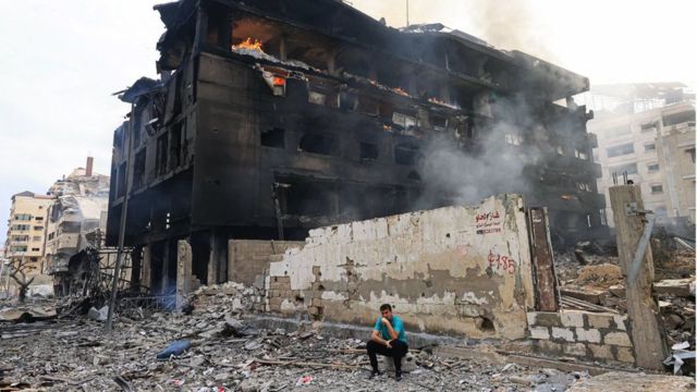 2023年10月10日，以色列对加沙市里马尔区发动空袭后，一名巴勒斯坦男子坐在一座烧焦的建筑物前，大楼内部正熊熊燃烧。