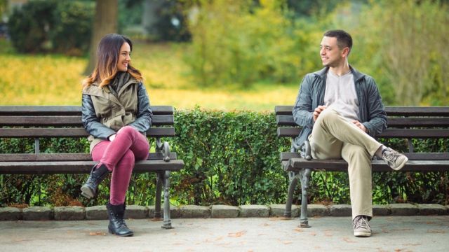 Un hombre y una mujer conversando sentados en dos bancas de un parque.