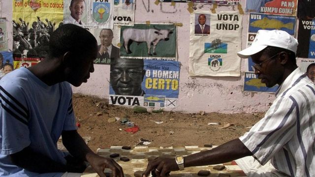Des résidents de Bissau s'adonnent au jeu de dame devant un mur couvert d'affiches de partis politiques (illustration)