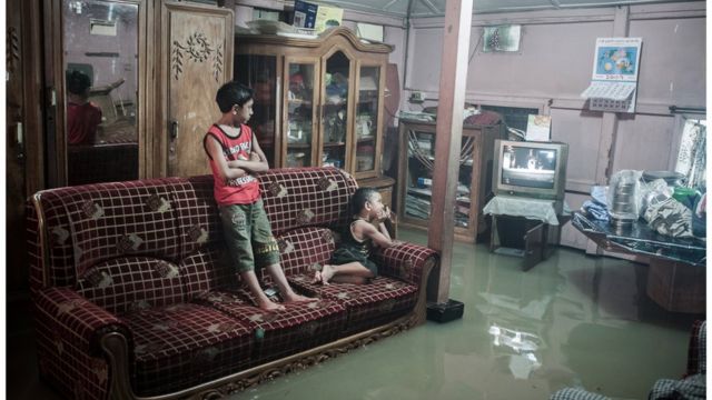 Crianças veem televisão em uma sala inundada