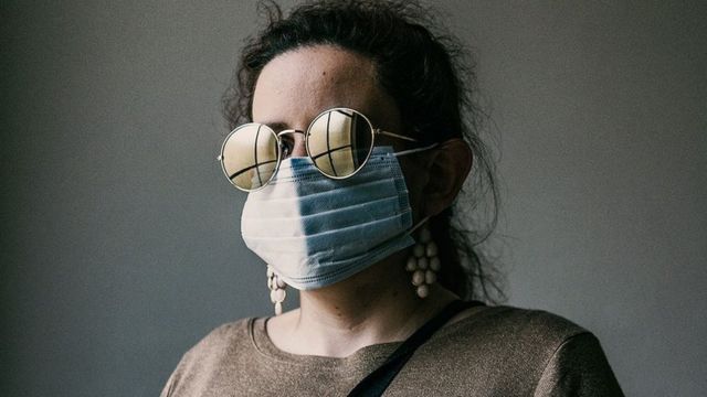 A blind woman stands with her walking cane whilst wearing a face mask and gloves