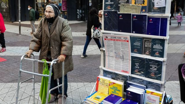 Una mujer mayor, con un andador, parada al lado de un quiosco que tiene ejemplares de la Constitución