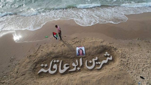 فلسطيني يرفع علم فلسطين بجوار صورة واسم شيرين أبو عاقلة الذي رسم بالرمل على شاطىء غزة
